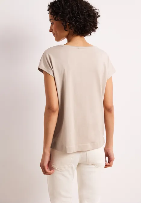 Person von hinten in beige T-Shirt und weißer Hose vor hellem Hintergrund.